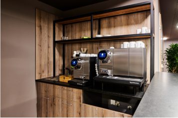 Ansicht eines silbernen Kaffeeautomaten auf einer Arbeitsfläche mit Tassen anderem Zubehör für die Kaffeezubereitung.